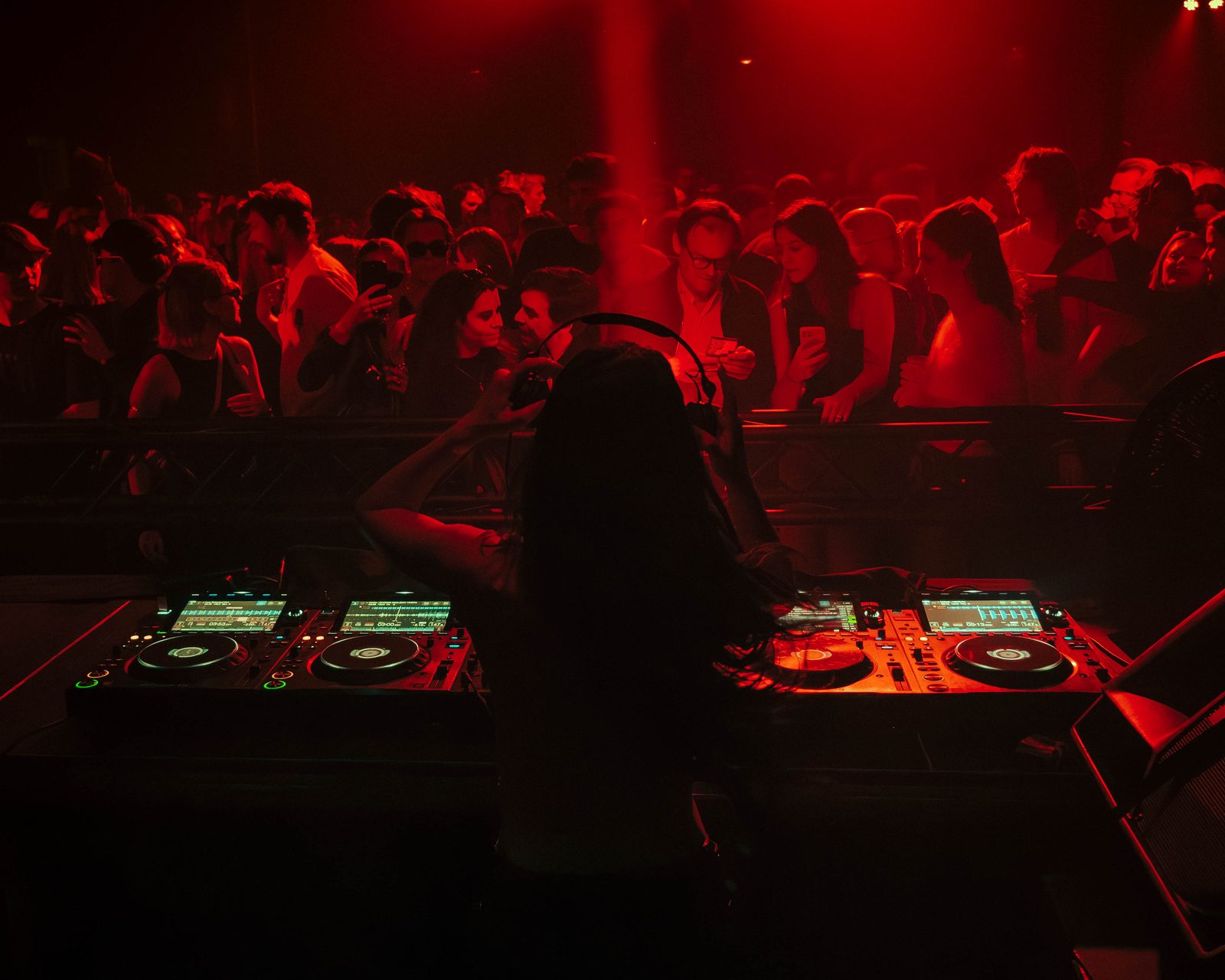 DJ silhouetted behind a dual CDJ booth with the crowd lit red beyond the barrier at a KompaanEvents night